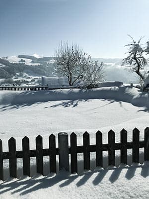 Dunkler Lattenzaun mit Schneehaube, dahinter der Blick über verschneite Wiesen