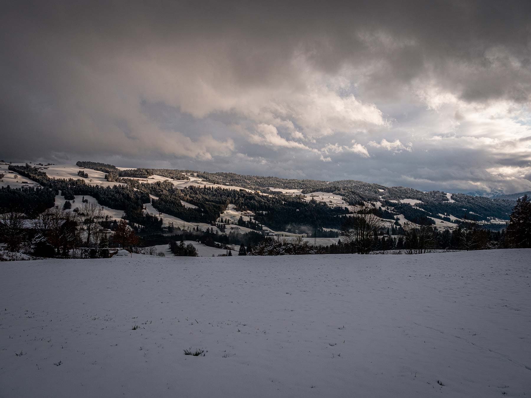 Verschneite Hügelkette unter schwerer Wolkendecke, im Vordergrund ein unberührtes Schneefeld