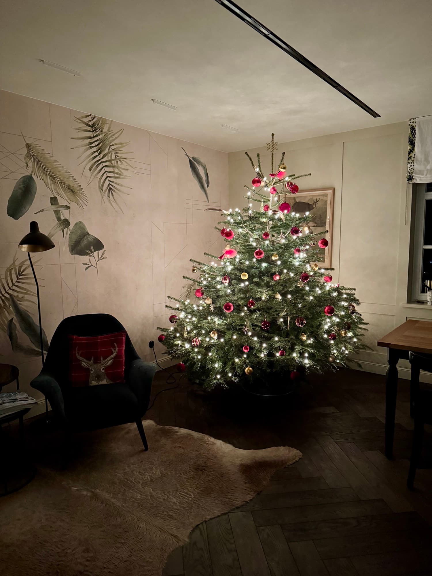 Geschmückter Christbaum mit warmen Lichtern und roten Kugeln im Aufenthaltsraum, daneben ein dunkler Sessel mit Hirschkissen auf einem Fell, im Hintergrund die Blättertapete