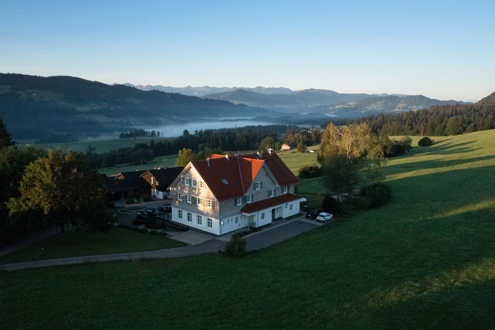 Luftaufnahme der Alpenloge im Morgenlicht: das Haus mit rotem Ziegeldach am Wiesenhang, dahinter Nebel im Tal und die Kette der Allgäuer Alpen