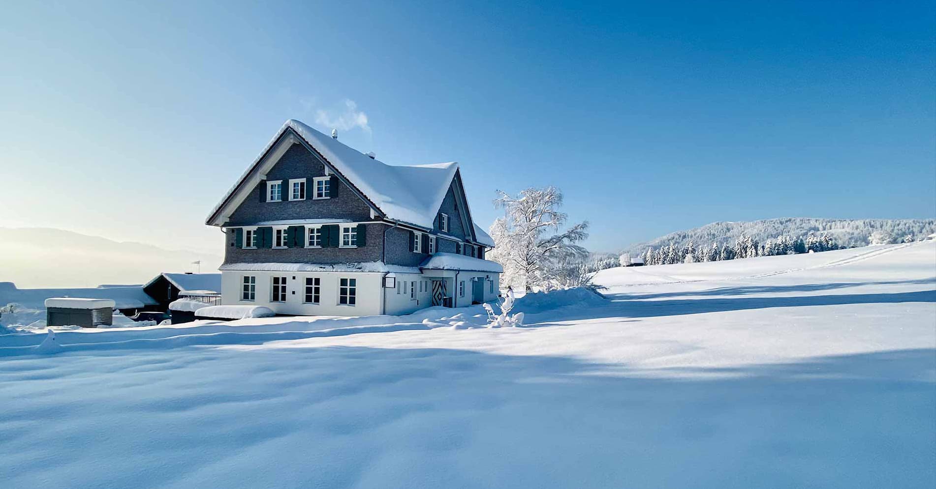 Die Alpenloge im Tiefschnee unter klarem blauem Himmel: das Haus mit verschneitem Satteldach und grünen Fensterläden, davor eine unberührte Schneefläche, rechts ein vereister Baum und bewaldete Hügel