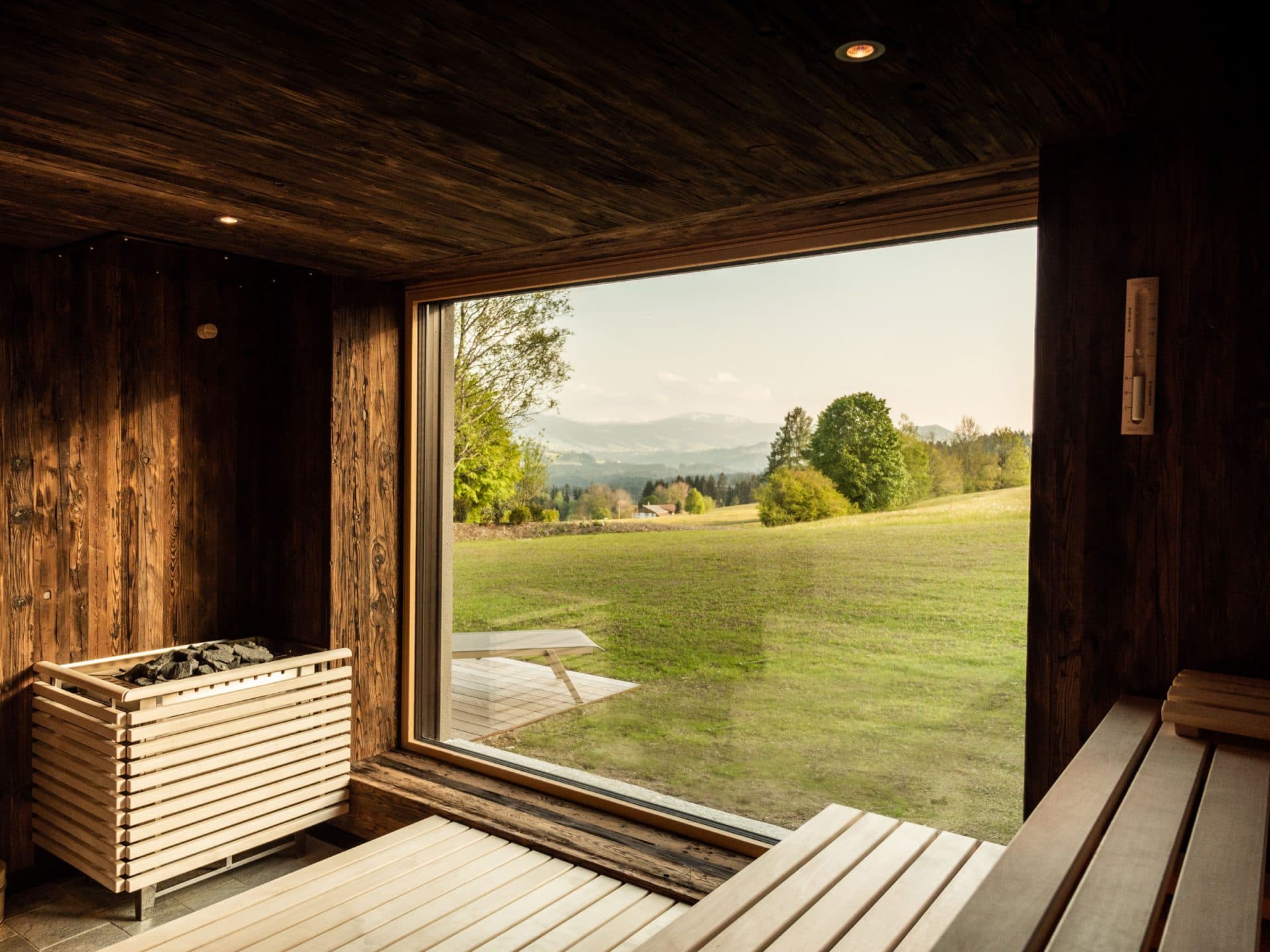 Panorama sauna in dark reclaimed wood, sauna stove with stones, floor-to-ceiling window onto the meadow and mountains beyond