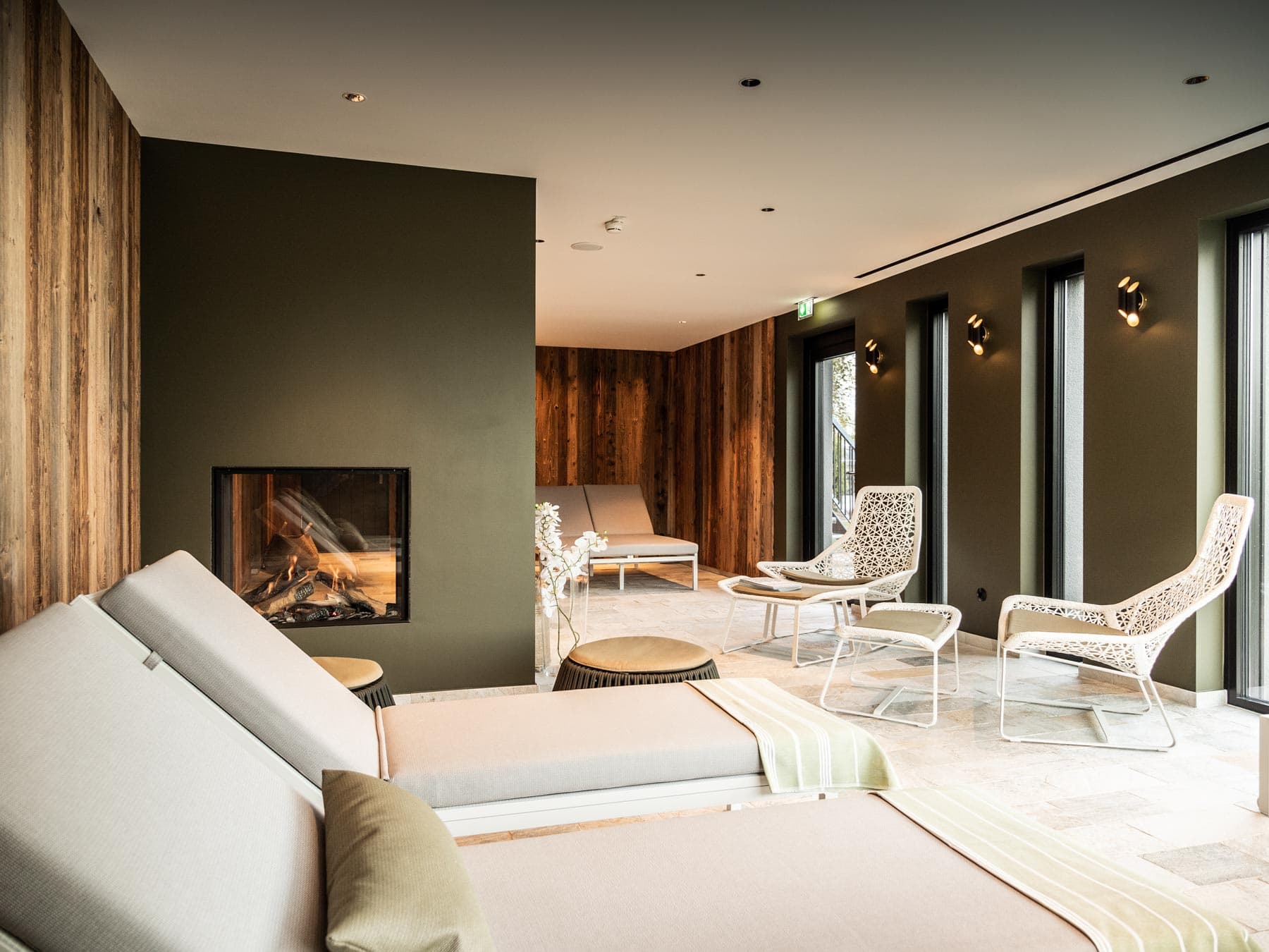 Relaxation room with a burning fireplace, light-coloured loungers and woven chairs against an olive-green wall and reclaimed wood