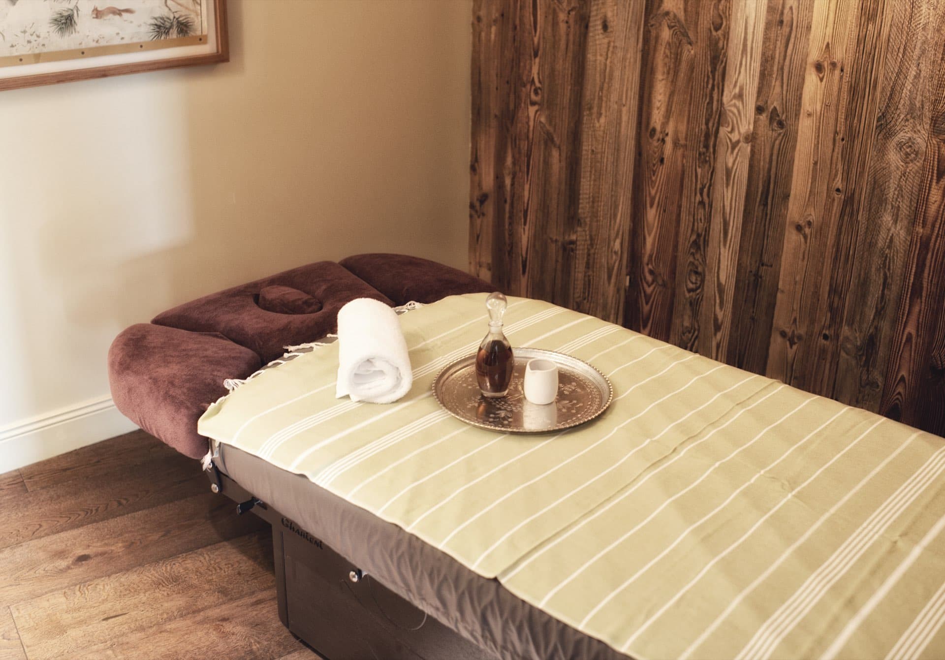 Massageliege mit gestreiftem Tuch, Handtuch und Ölkaraffe auf einem Messingtablett vor einer Altholzwand