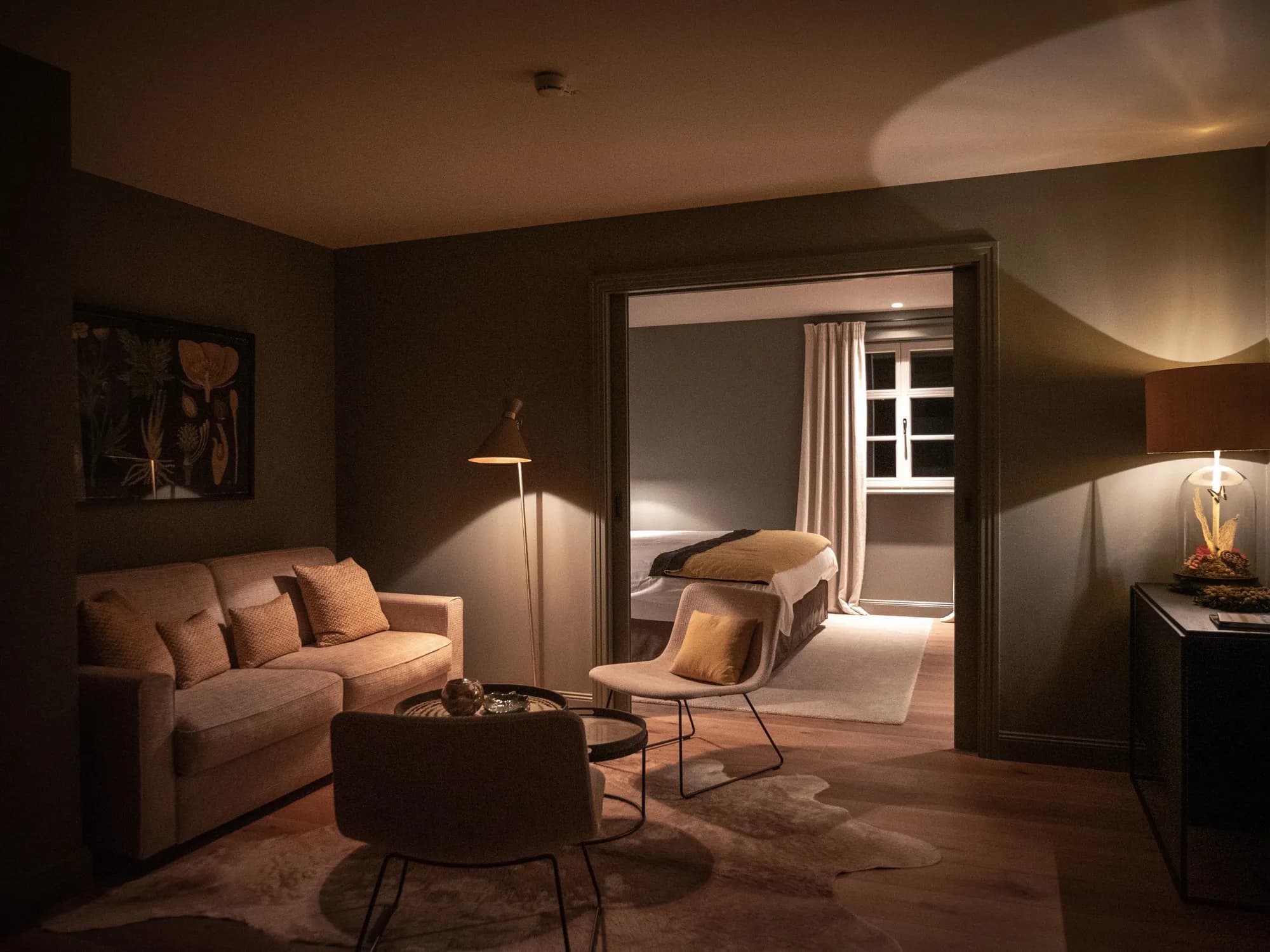 Wohnbereich der Suite Drei am Abend, Sofa und Stehlampe, Blick durch die offene Tür ins Schlafzimmer