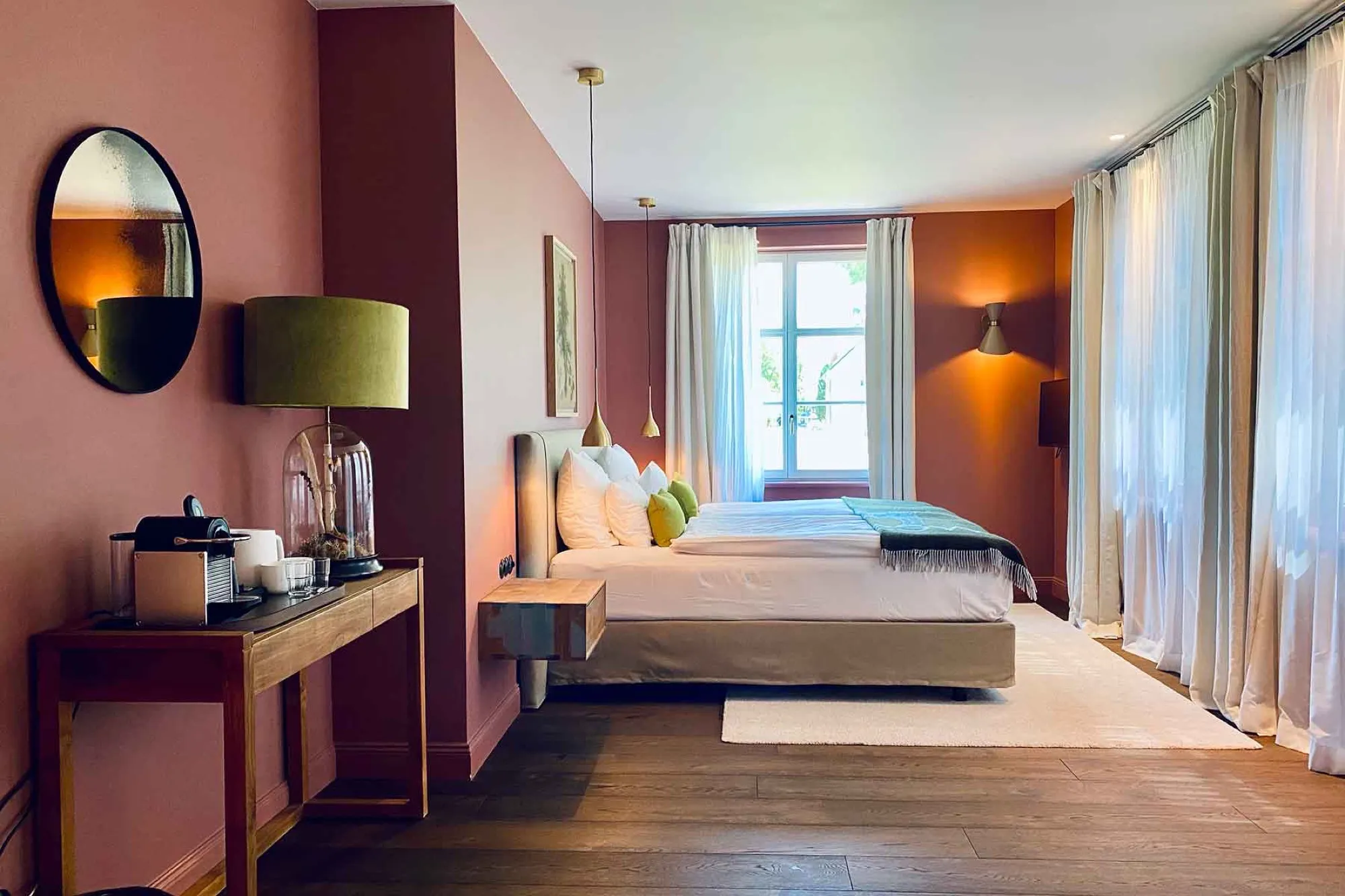 Overview of Suite One with the bed against a pink wall, a console table with a round mirror, tall curtained windows